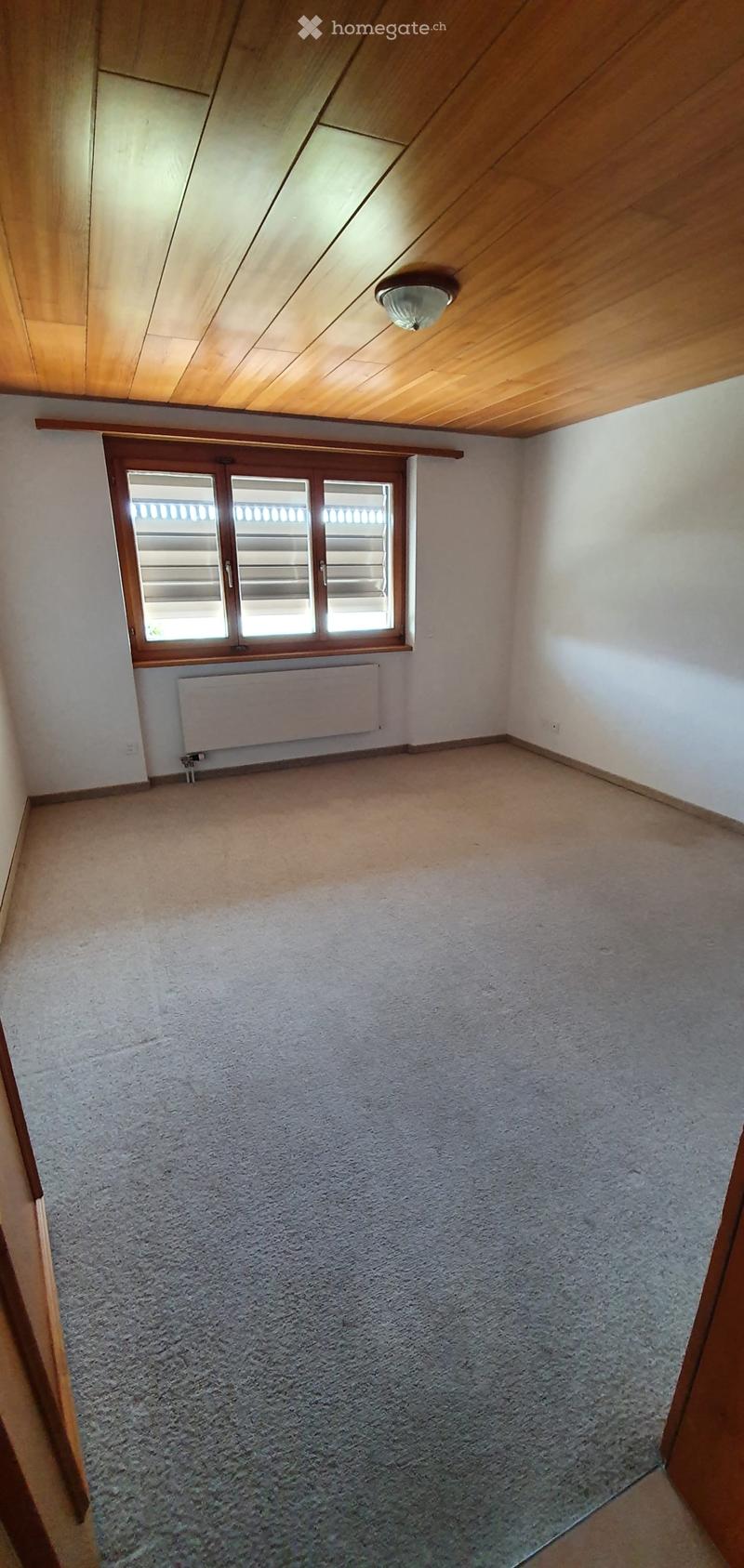 Empty room with wooden slanted ceiling, carpeted floor, white walls, and a window with blinds. A radiator is visible below the window.