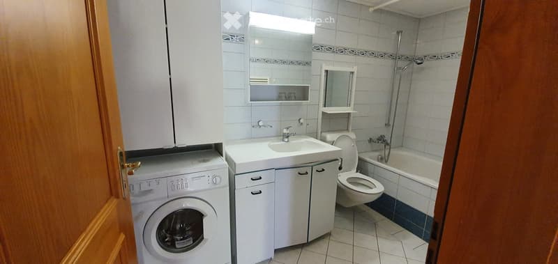 Bathroom with a white sink, mirror, toilet, bathtub, and a washing machine. The walls are tiled, and there is a wooden door frame visible.