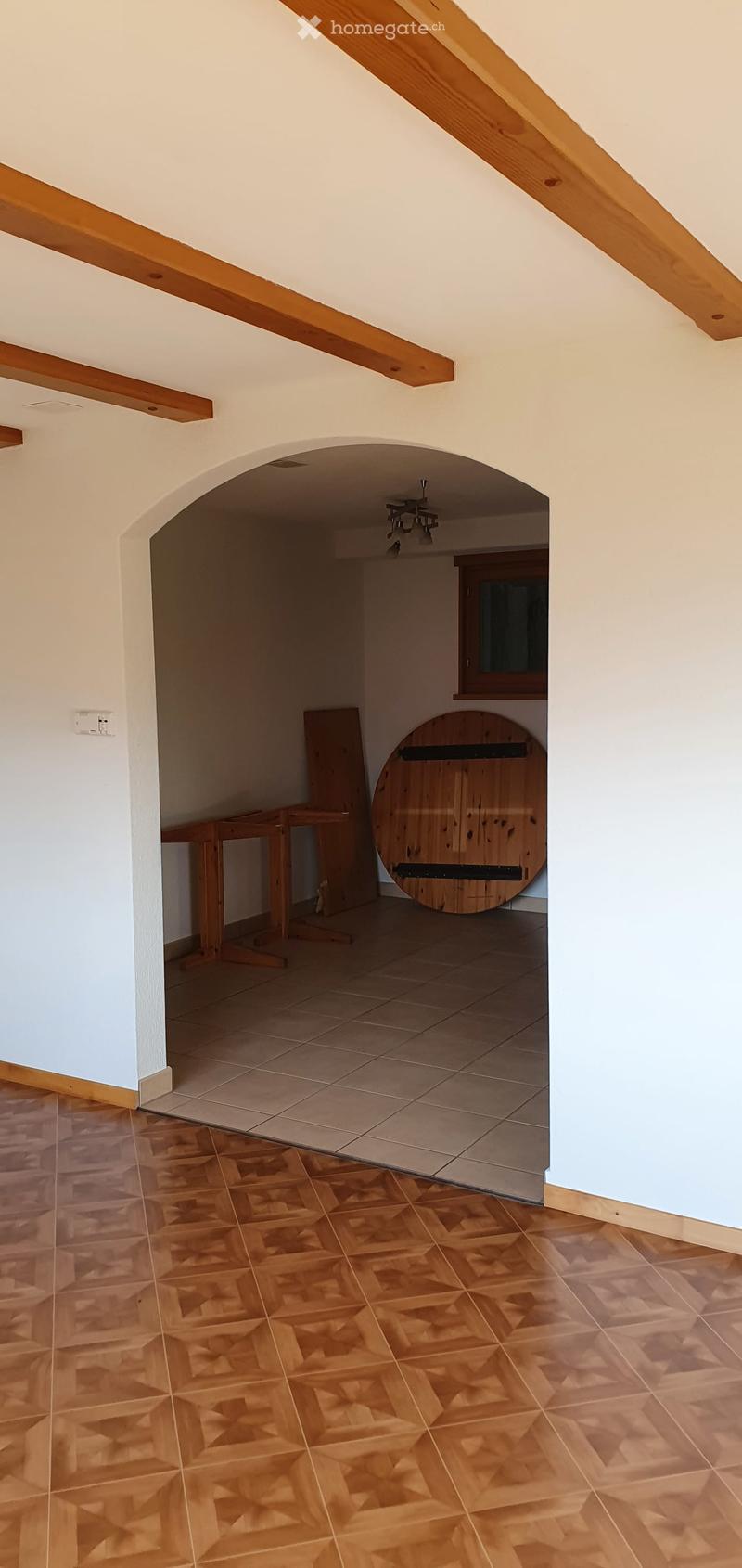 A narrow hallway with wooden beams on the ceiling, light-colored walls, and a mix of tiled and wooden flooring. An arched doorway leads to another room with a small table and a round wooden object.