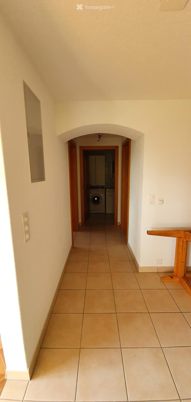 Empty hallway with tiled floors, white walls, and a small wooden table on the right side. A doorway leads to a room with a washing machine and dryer visible in the background.