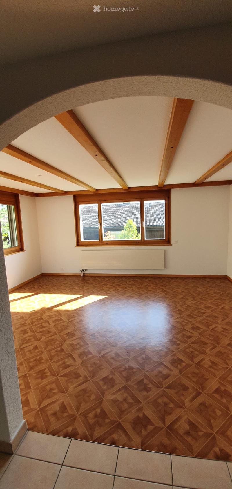 Empty attic room with wooden floor, exposed wooden beams, large windows, and radiator.