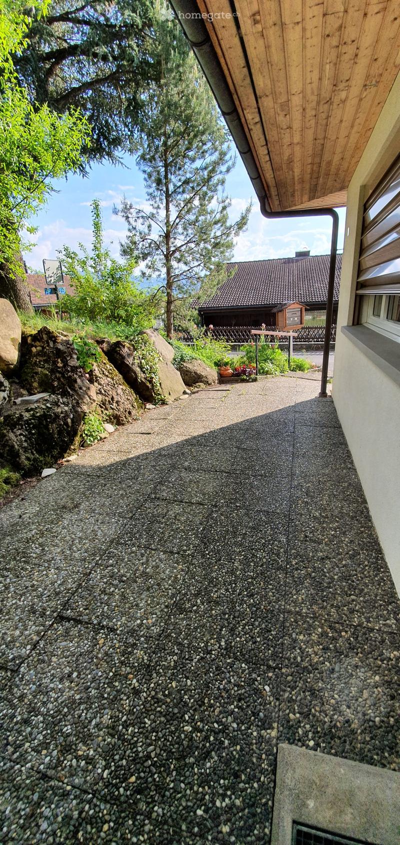 Gravel pathway leading up to a house with a wooden roof and white walls, surrounded by greenery and trees.
