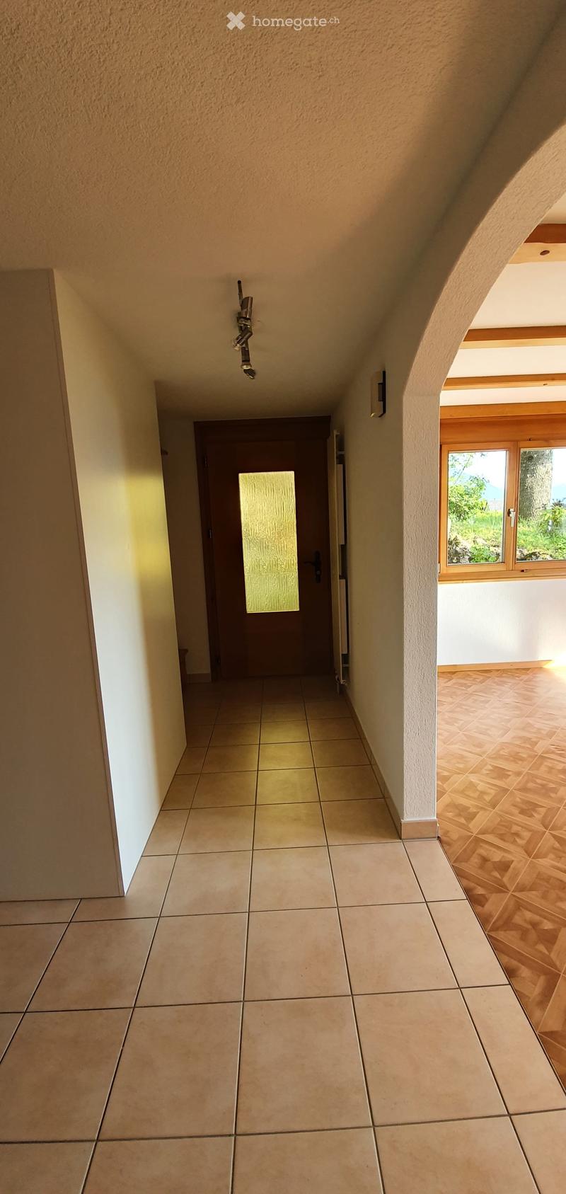 Empty hallway with tiled floor, beige walls, and a wooden door at the end. A window on the right side shows a view of greenery outside.