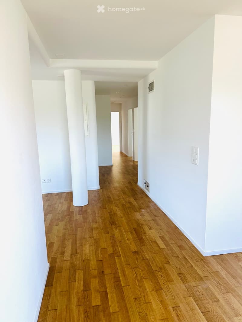 Empty hallway with wooden flooring, white walls, and a central column. Natural light filters through a window at the far end.