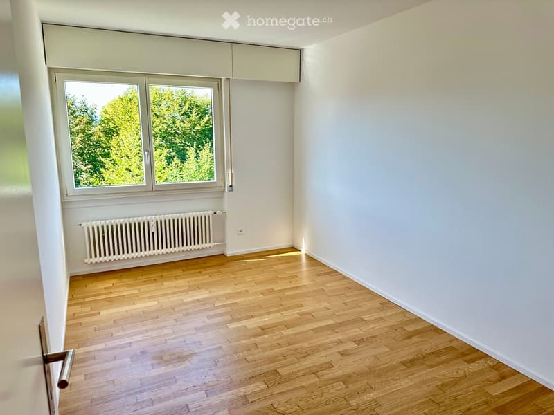 Empty room with light wooden flooring, white walls, a large window with a view of greenery, and a radiator below the window.