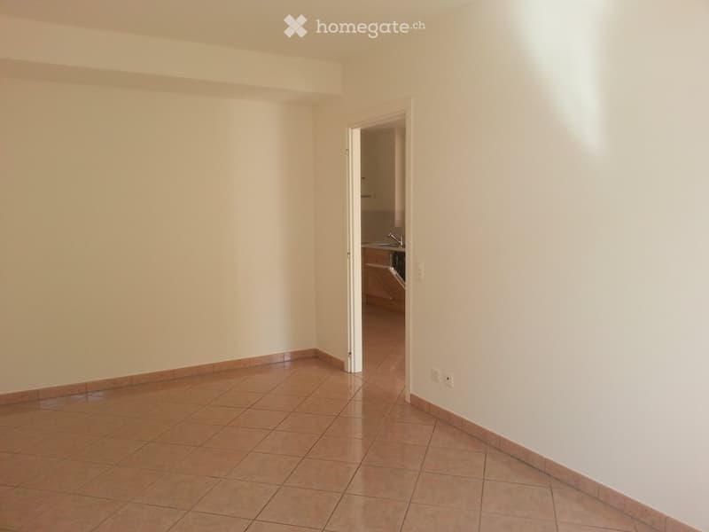 Empty room with beige tiles and white walls, open door leading to kitchen with sink and cabinets.