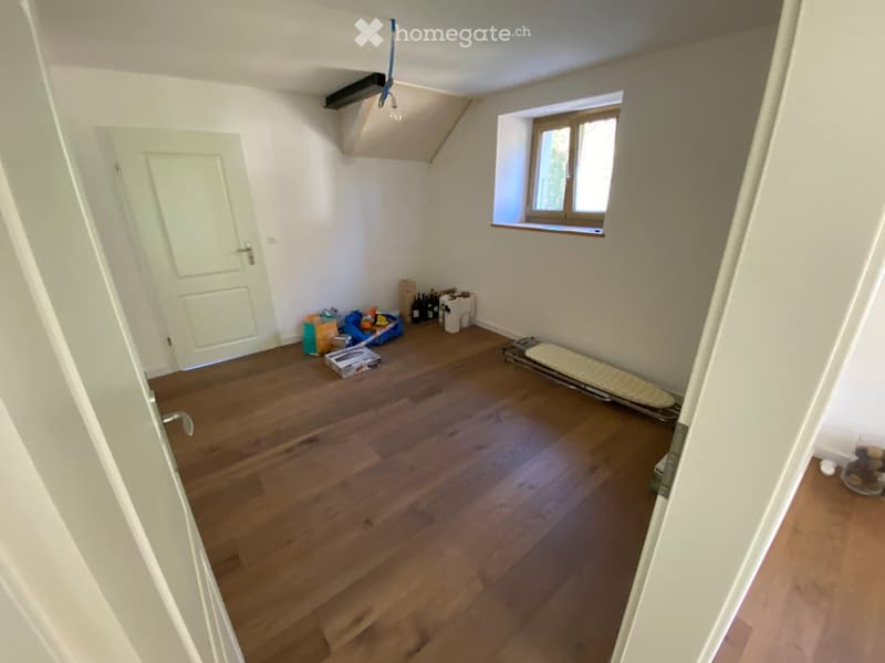 Empty room with wooden flooring, white walls, a window, and a door. Some storage items are visible near the window.