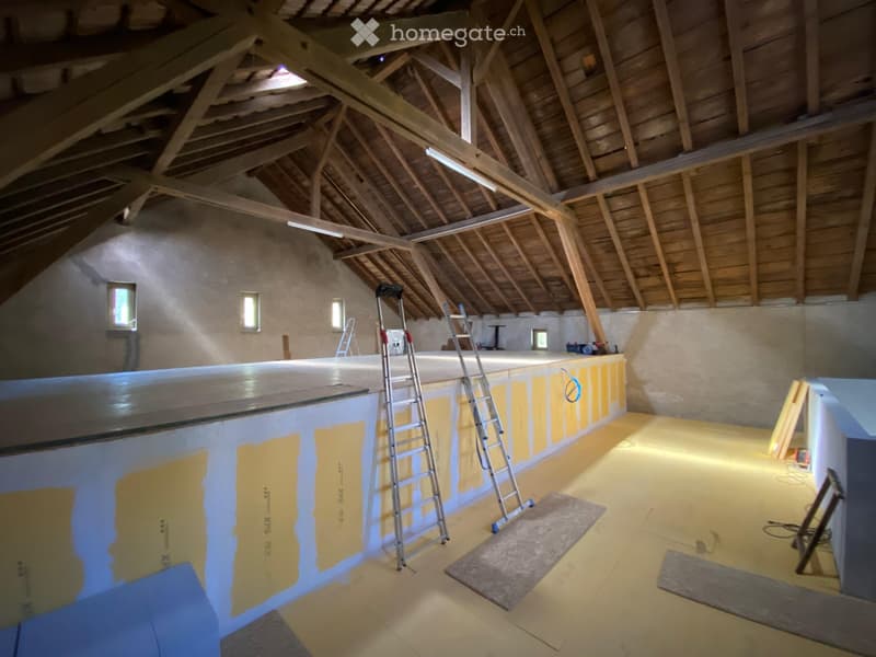 Empty attic space with exposed wooden beams, unfinished walls, and scattered construction materials including ladders and boards.