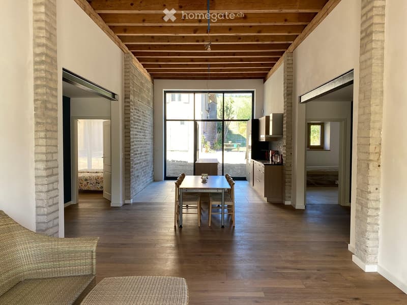 Open-plan living and dining area with wooden flooring, exposed wooden ceiling beams, brick walls, and large glass doors leading to an outdoor space. Visible furniture includes a sofa, dining table with chairs, and kitchen cabinets.