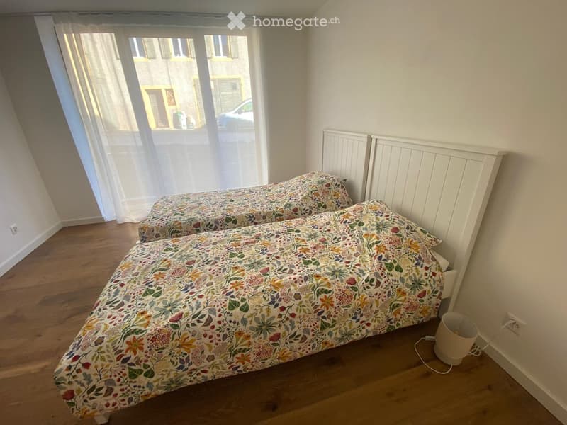 Cozy bedroom with two single beds, floral bedding, wooden flooring, radiator, and large window with white curtains.