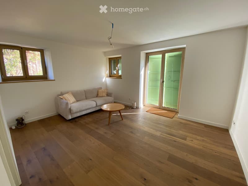 Bright living room with wooden flooring, a grey sofa with throw pillows, a round wooden coffee table, and large windows with a sliding glass door leading outside.