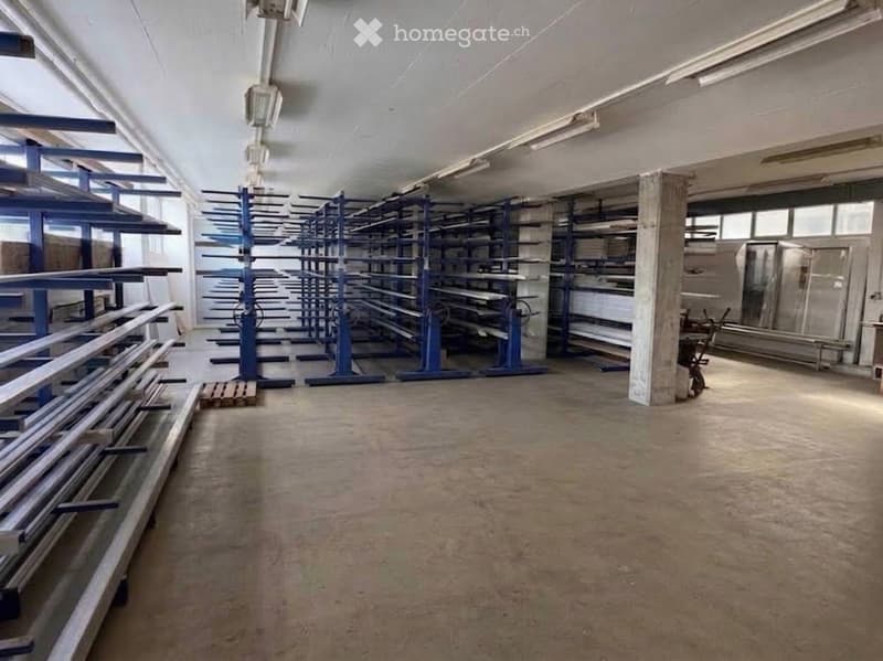 Empty warehouse space with concrete floor, metal shelving units, and large windows allowing natural light. No furniture or equipment present.