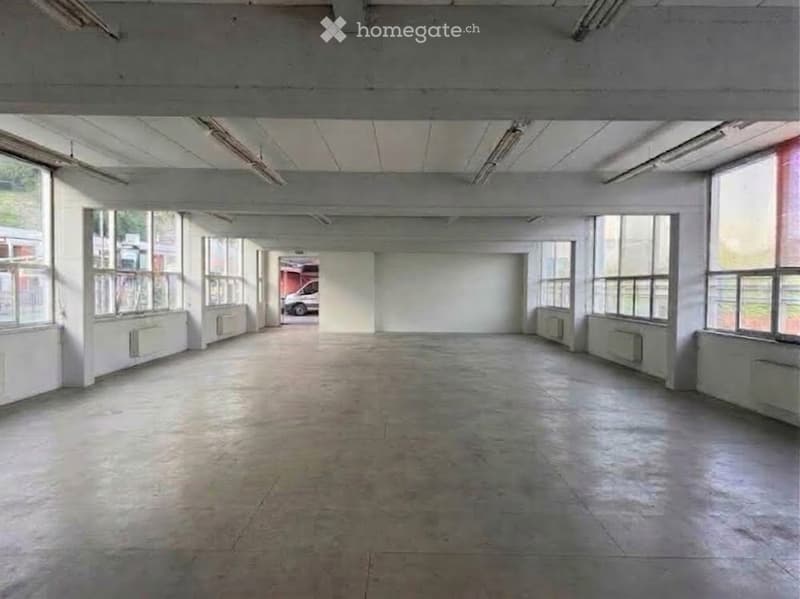 Large, empty industrial-style room with concrete floors, white walls, and large windows allowing natural light. Exposed ceiling beams and fluorescent lighting fixtures are visible.