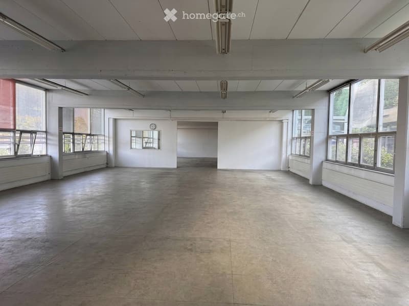 Large, empty industrial-style room with concrete floors, white walls, large windows, and exposed ceiling beams. No furniture or equipment present.