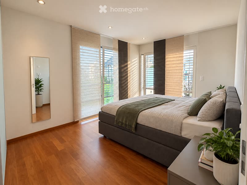 Modern bedroom with wooden flooring, large windows with blinds, a grey upholstered bed with pillows and a green throw, a nightstand with a potted plant, and a mirror on the wall.