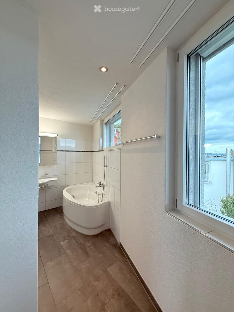 Modern bathroom with a freestanding bathtub, sink, mirror, and cabinet. Large windows offer natural light and a view.