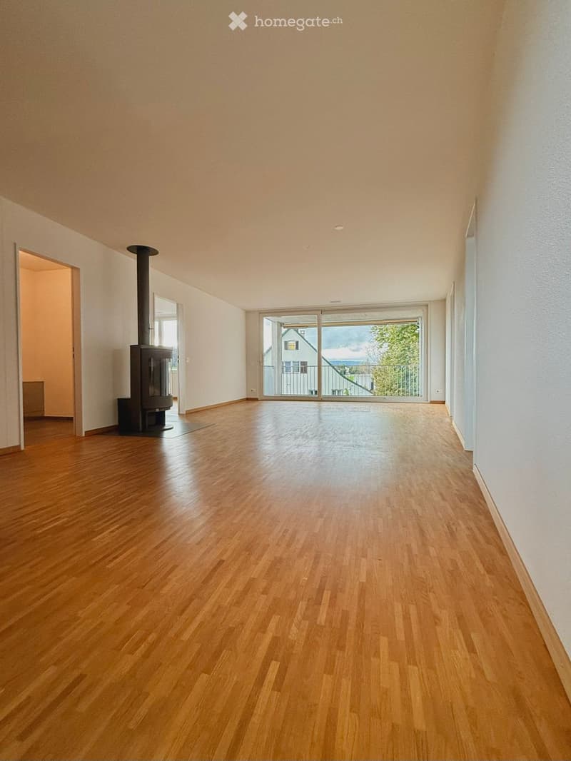 Empty room with wooden flooring, white walls, and a large sliding glass door leading to a balcony. A wood-burning stove is visible on the left side.