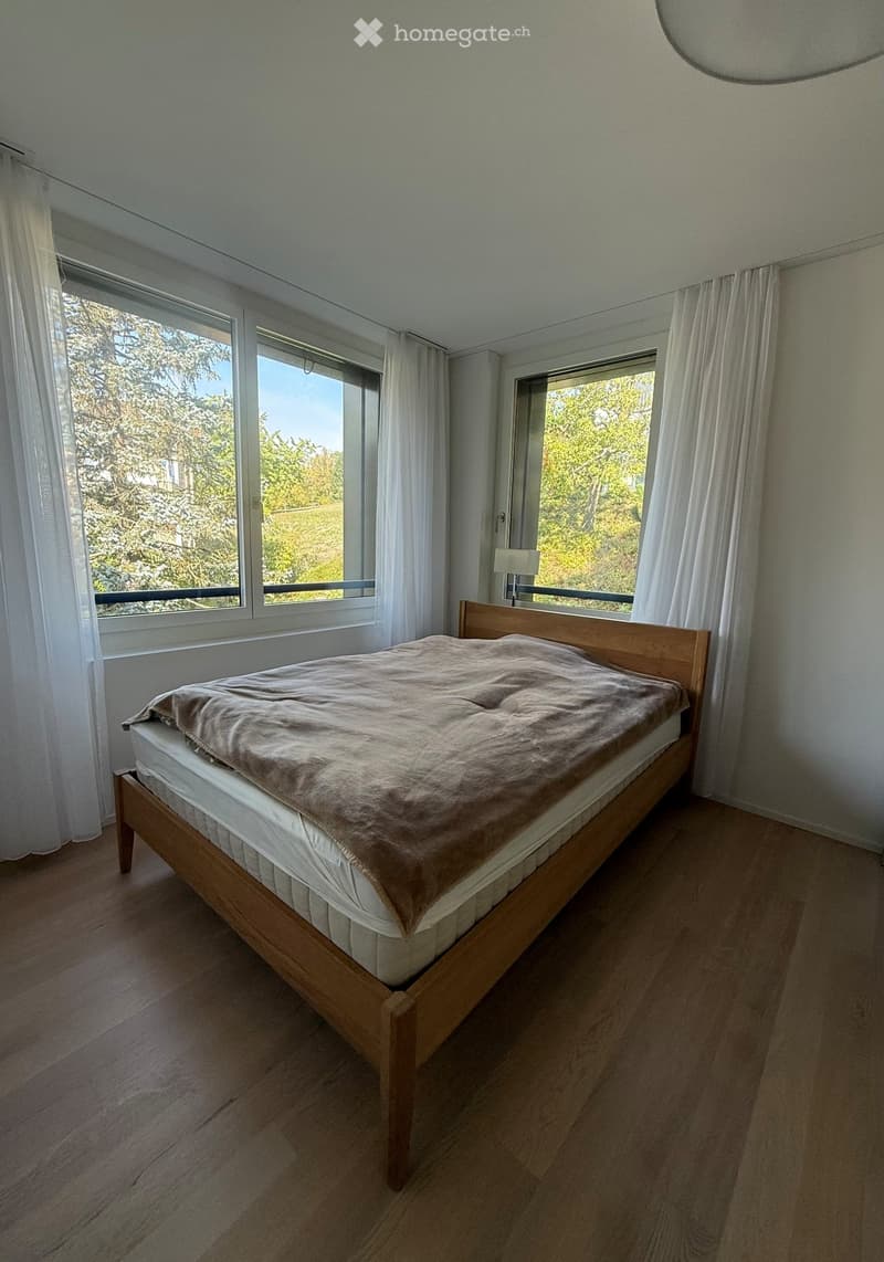Bright bedroom with wooden floor, large windows with white curtains, and a single wooden bed with beige bedding.