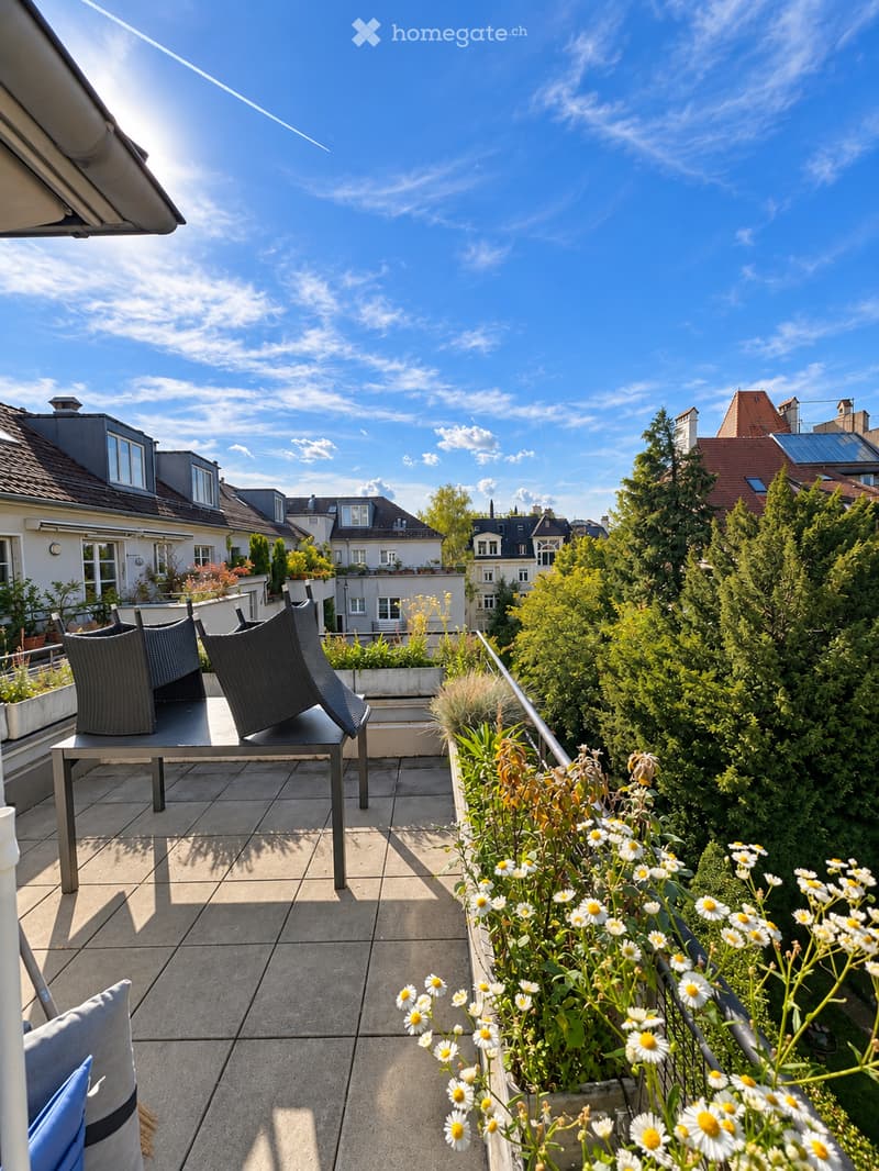 A rooftop terrace with a tiled floor, a metal table, two black wicker chairs, and a small garden bed with white daisies.