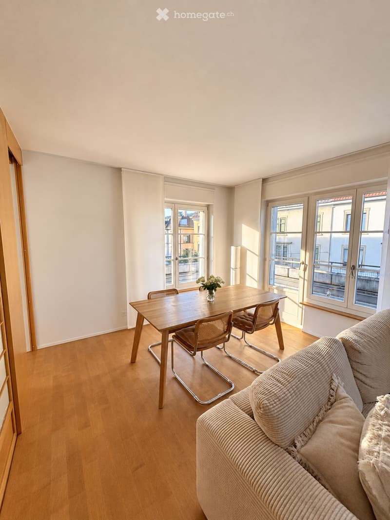 Bright, open living and dining area with wooden flooring, large windows, a sofa, dining table with chairs, and a vase of flowers.