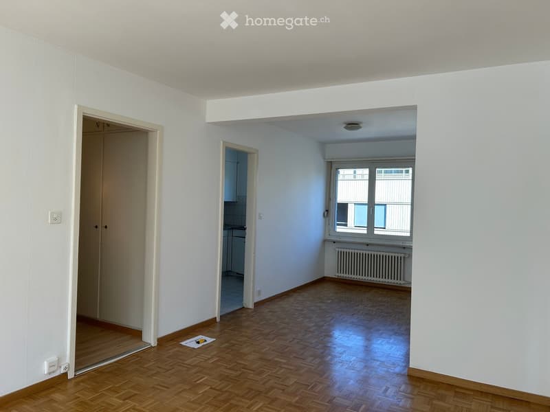 Empty room with wooden flooring, white walls, and large windows. Adjacent rooms visible through open doorways.