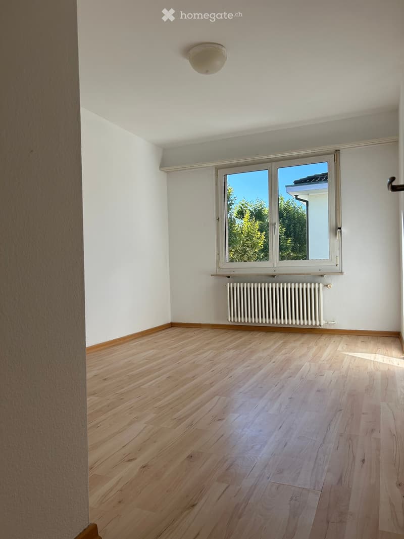 Empty room with light wooden flooring, white walls, a large window with a view of trees, and a radiator below the window.