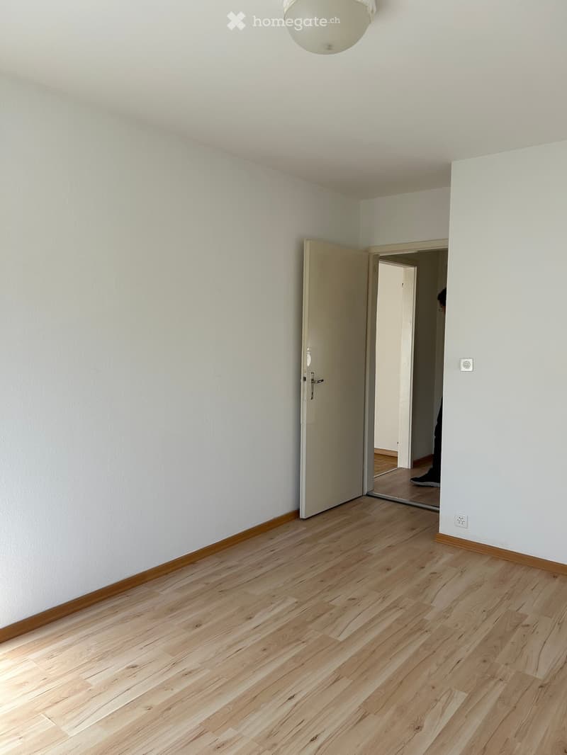 Empty hallway with light wooden flooring, white walls, and a single open door leading to another room.