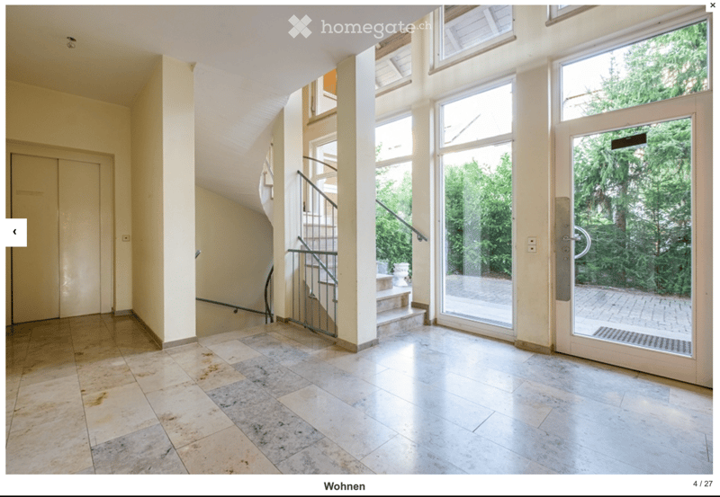 Bright hallway with tiled flooring, large windows, and a staircase leading upwards. Features include double doors, a metal railing, and a view of greenery outside.