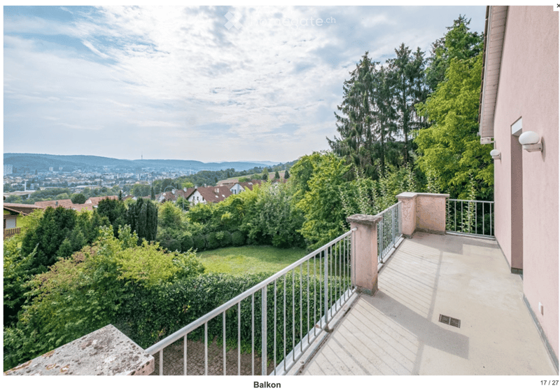 Balcony with metal railing, stone balusters, and a view of a green garden and cityscape beyond.