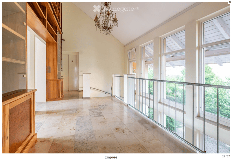 Bright, spacious hallway with high ceilings, large windows, and a chandelier. Features a wooden cabinet on the left and a staircase leading upwards.