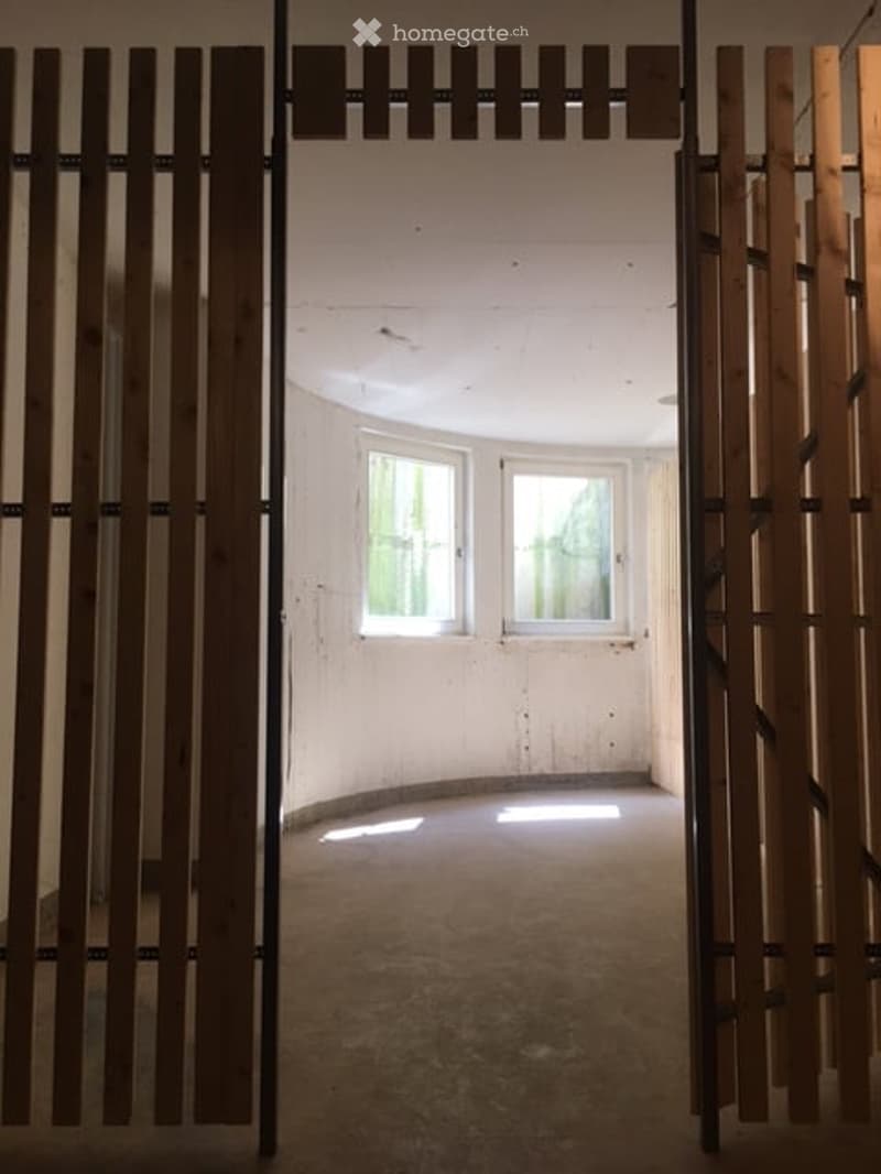 Empty room with exposed concrete floor, white walls, and two windows allowing natural light. The space is unfurnished and appears to be a simple, open area.