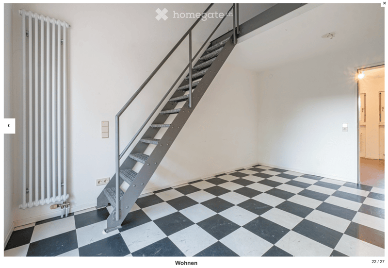 Empty room with black and white checkered floor, white walls, metal staircase leading upwards, radiator on left wall, and a doorway to another room on the right.