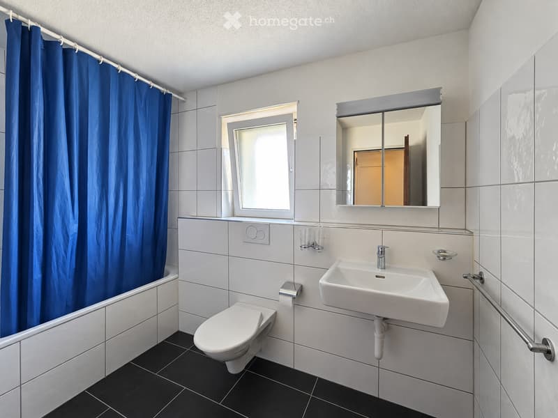 Modern bathroom with white tiles, a toilet, a sink, a mirror, and a bathtub with a blue curtain.