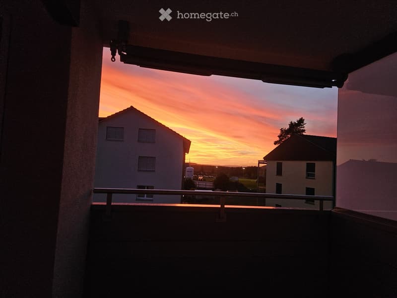 Balcony overlooking a residential area during sunset, with a railing and distant buildings visible.