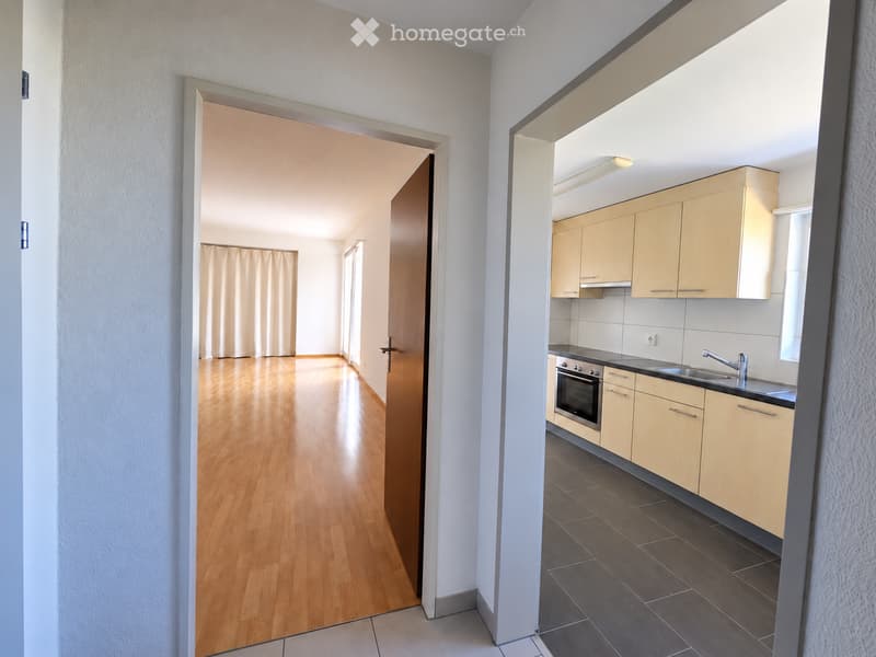 Empty room with wooden flooring, white walls, and a doorway leading to another room. The adjacent kitchen features light wood cabinets, a sink, and an oven.