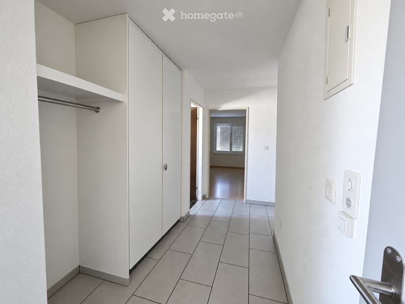 Clean, modern hallway with white walls, tiled floor, built-in closet with shelves and hanging rod, and a door leading to another room with wooden flooring and a window.