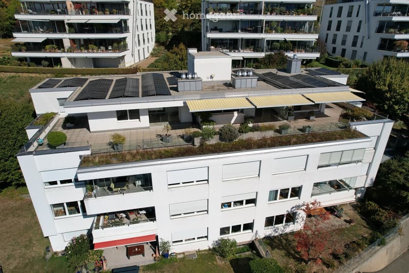 Aerial view of a modern, multi-story residential building with white exterior, large windows, balconies, and rooftop terraces. The building features solar panels on the roof and is surrounded by greenery and neighboring structures.