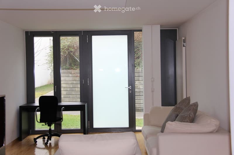 A modern living room with a white sofa, ottoman, desk, and chair, featuring large sliding glass doors leading to an outdoor area.