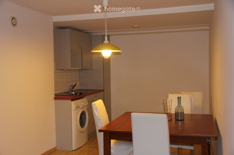 A modern, compact kitchen and dining area with a wooden dining table, white chairs, a washing machine, and a small sink area with a faucet. The space features light-colored cabinets and a hanging pendant light.