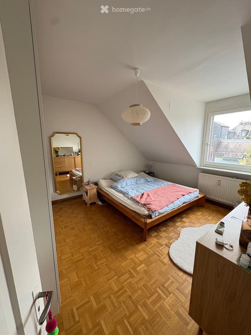 Cozy attic bedroom with wooden flooring, slanted ceilings, and large window offering a view. Features a single bed with blue and pink bedding, a nightstand, a mirror, and a small table with decorative items.