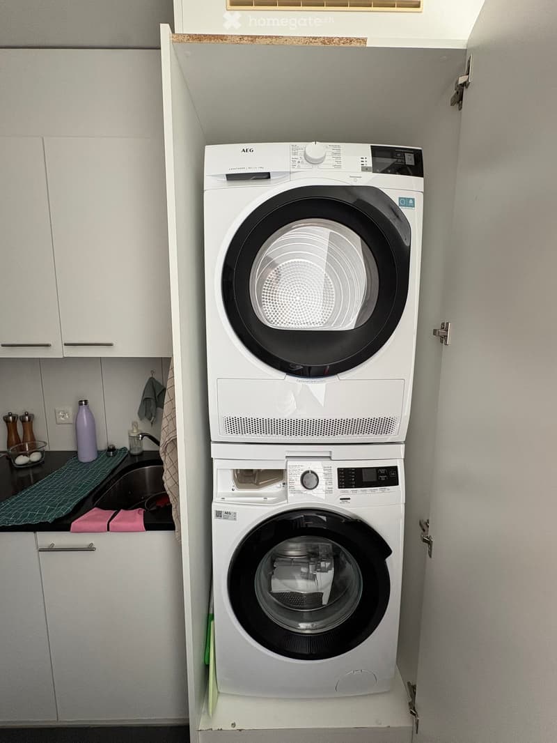 A compact bathroom with a stacked washing machine and dryer unit, a sink with a faucet, and various small items on the counter.