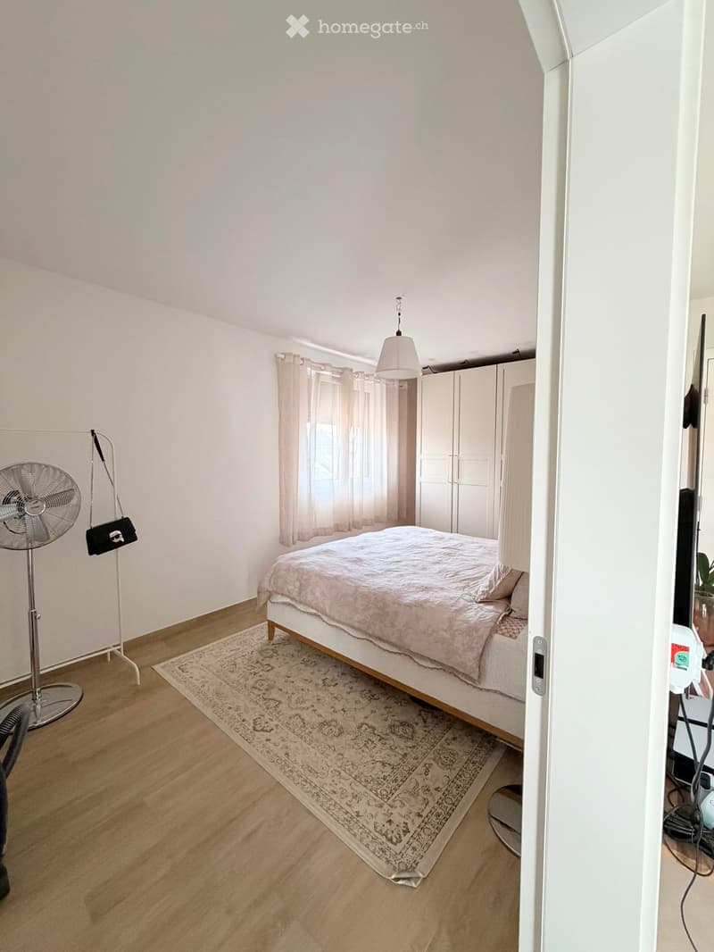 Bright bedroom with wooden flooring, a bed with a pink duvet, a large rug, a standing fan, and a built-in wardrobe.