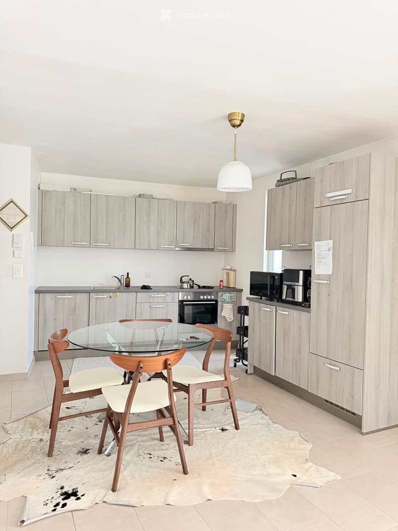 Modern kitchen with light wood cabinets, stainless steel appliances, a glass dining table with four wooden chairs, and a pendant light fixture.