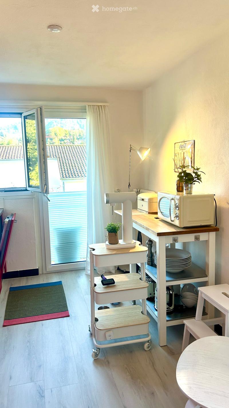 A small, well-lit kitchen area with a microwave, a small table, a stool, a rolling cart, a lamp, and potted plants. The room features light wooden flooring and a large window with blinds that opens to a view of a tiled roof and greenery.