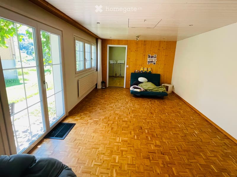 A spacious, well-lit room with wooden flooring, large windows, and a sofa with a green blanket. There is a small table with decorative items and a stool nearby.