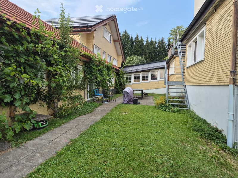 A small, enclosed garden area with a concrete pathway, grassy lawn, and a few outdoor chairs and a table. The garden is bordered by residential buildings with yellow facades and red-tiled roofs.