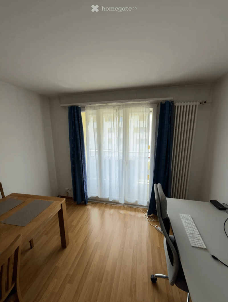 A small, unfurnished room with wooden flooring, a desk with a chair, a small dining table with chairs, and a window with curtains.