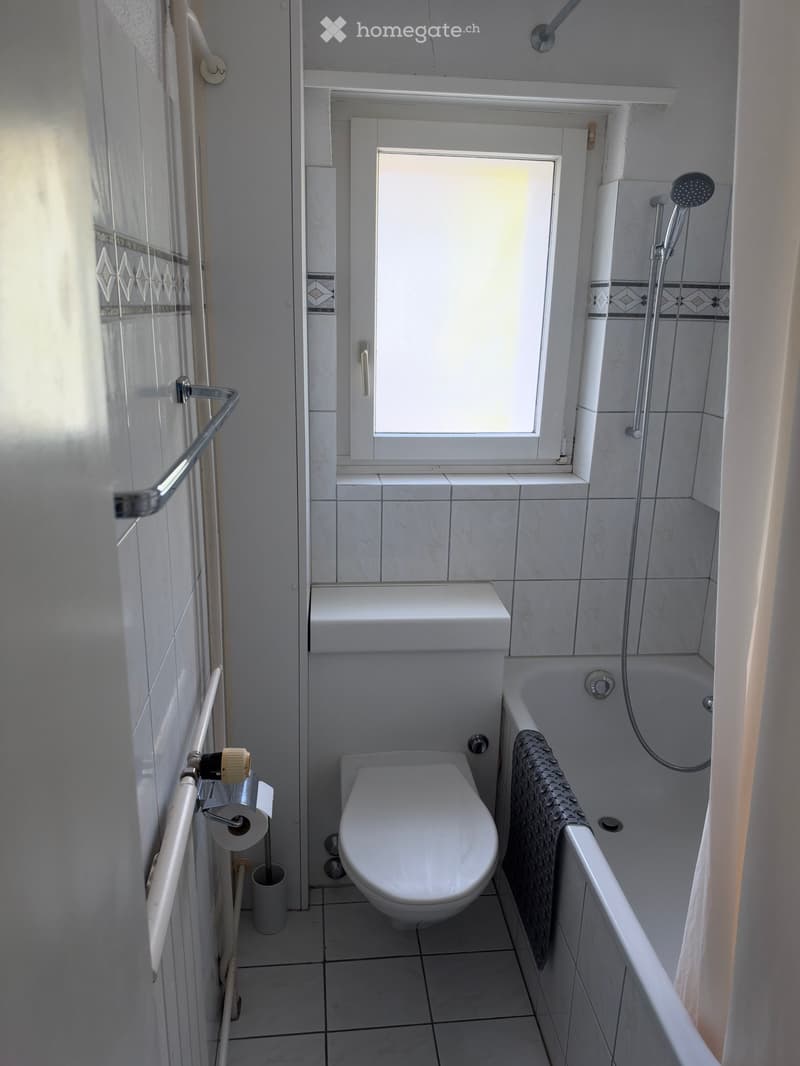 Modern bathroom with white tiles, a toilet, bathtub, shower, towel, and toilet paper holder.