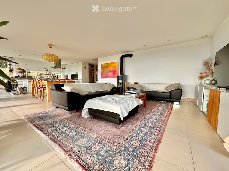 Modern living room with a large sectional sofa, coffee table, ottoman, and fireplace. The space features a colorful rug, wooden chairs, and a kitchen area visible in the background.