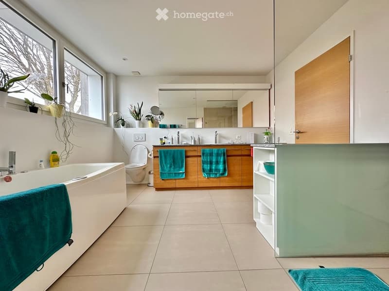 Modern bathroom with a bathtub, toilet, sink, large mirror, wooden cabinets, and green towels. Features large windows with plants on the windowsill and a shelf with decorative items.